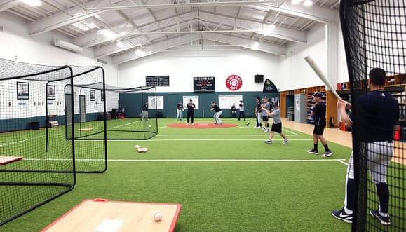 An indoor baseball clinic facility setup featuring a spacious training area filled with various equipment. In the foreground, there are batting cages with safety nets and baseballs scattered around, alongside training aids like pitching mounds and strike zone posters. The middle ground showcases youth baseball players, dressed in professional athletic attire, actively practicing their swings and pitch techniques with coaches demonstrating drills. The background reveals high ceilings with bright, soft lighting, showcasing wall-mounted scoreboard and storage for baseball gear. The atmosphere is energetic and focused, emphasizing the importance of practice and development in a well-equipped facility. The image is captured at a wide-angle perspective to provide an expansive view of the entire setup. An indoor baseball clinic facility setup featuring a spacious training area filled with various equipment. In the foreground, there are batting cages with safety nets and baseballs scattered around, alongside training aids like pitching mounds and strike zone posters. The middle ground showcases youth baseball players, dressed in professional athletic attire, actively practicing their swings and pitch techniques with coaches demonstrating drills. The background reveals high ceilings with bright, soft lighting, showcasing wall-mounted scoreboard and storage for baseball gear. The atmosphere is energetic and focused, emphasizing the importance of practice and development in a well-equipped facility. The image is captured at a wide-angle perspective to provide an expansive view of the entire setup.