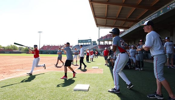 A dynamic baseball training scene showcasing "station rotations" at a preseason jamboree. In the foreground, a diverse group of players in modest athletic wear engage in different skill drills: a player practicing batting with a coach demonstrating techniques, another focusing on defensive maneuvers, and a third mastering baserunning. The middle ground features various training stations equipped with baseball equipment, such as bats, gloves, and bases, arranged effectively. In the background, a bright, clear sky illuminates a well-maintained baseball field, with spectators watching from a distance, creating an energetic yet focused atmosphere. The composition uses a wide-angle lens to capture the action and excitement, highlighting teamwork and skill development, while ensuring a vibrant and motivational mood pervades the scene.